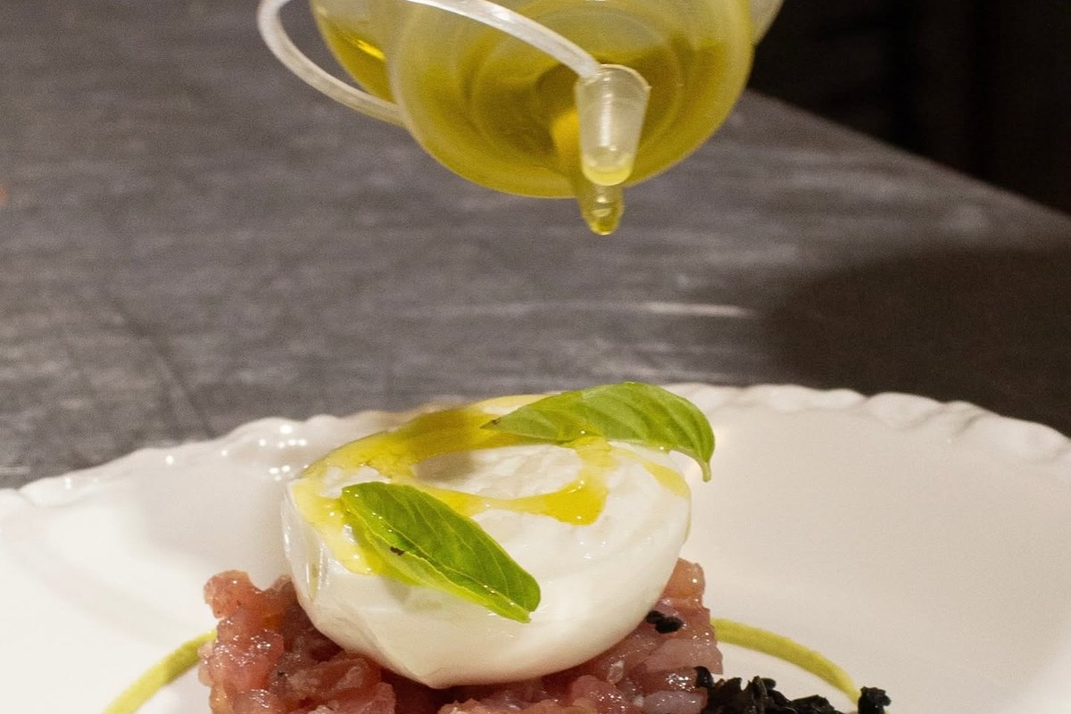 Olio dell'Agriturismo Abbatia versato da un'ampolla di vetro su burrata con basilico sopra una tartare, in piatto bianco