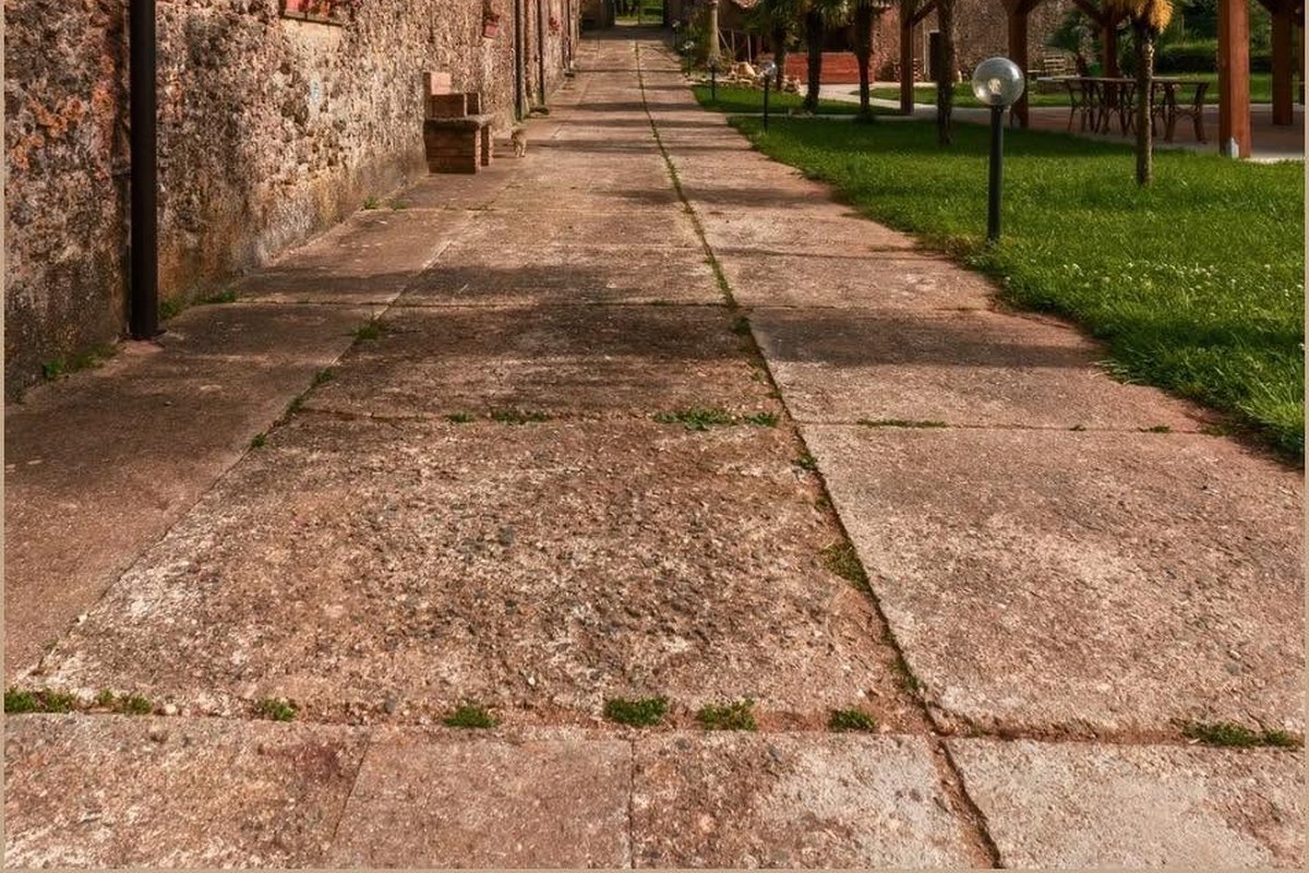 Vialetto lastricato in pietra lungo un muro rustico, con prato, lampioni a globo e pergolato di legno sullo sfondo